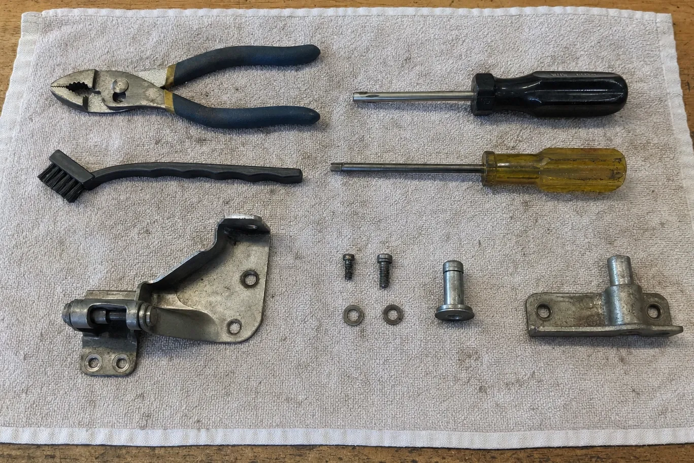 Pliers, a wire brush and two screwdrivers laid on a towel beside a dismantled door hinge and its screws